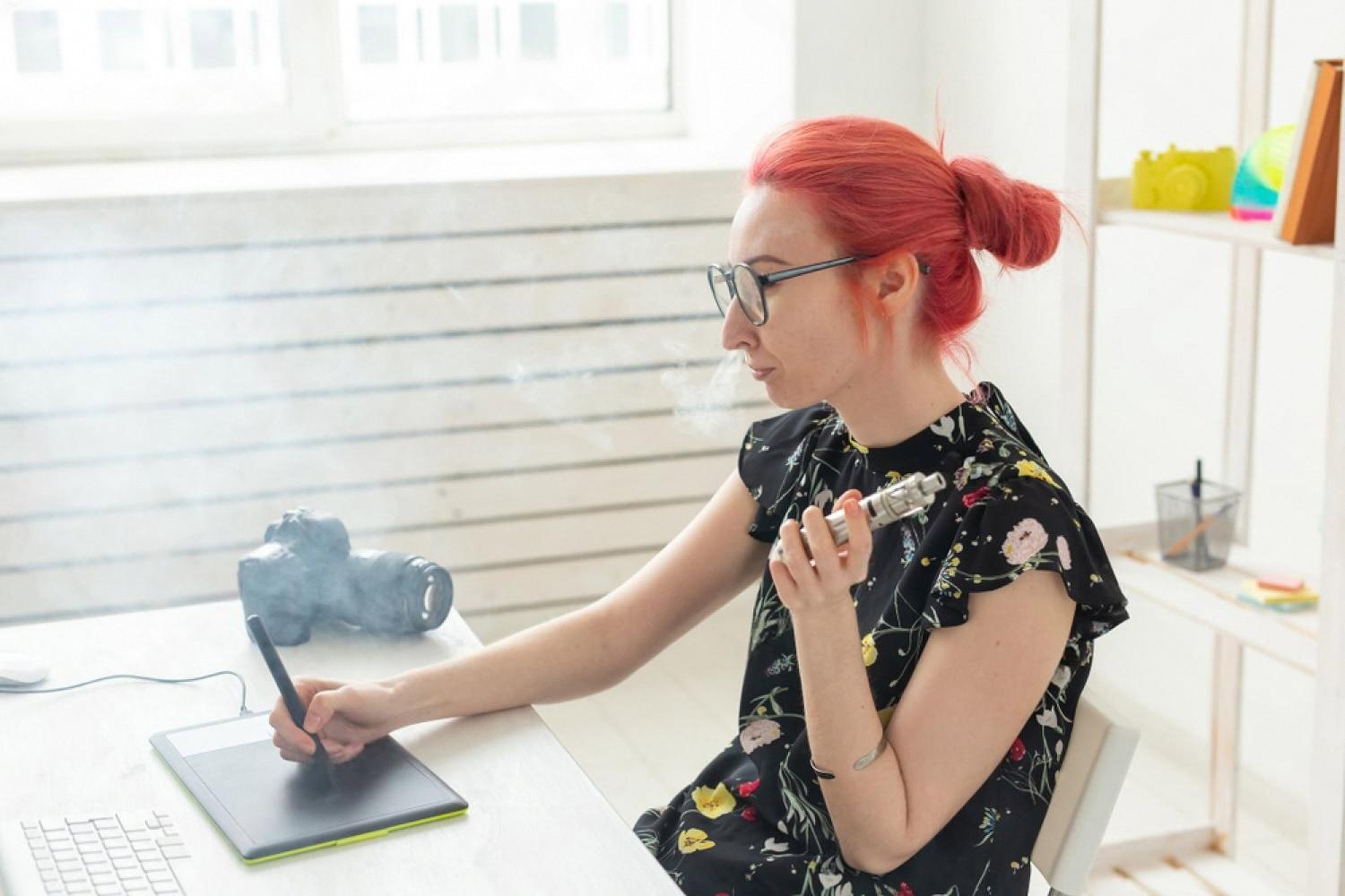 Young woman vaping at her desk while working, in a bright home office